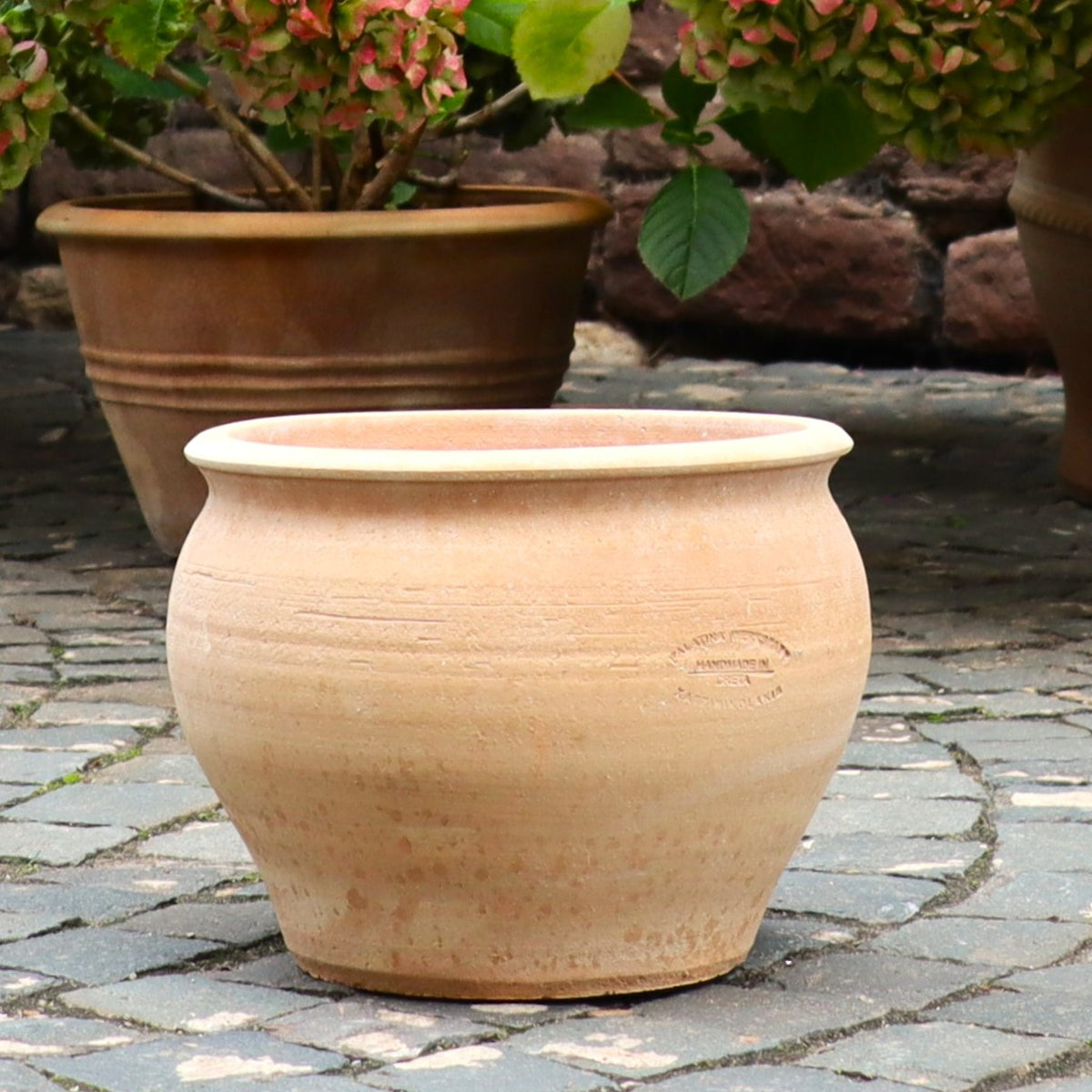 Ein runder, beiger Keramiktopf "Veronica" (D=30cm) für Pflanzen steht auf einem Kopfsteinpflaster im Freien, mit einem weiteren Pflanzentopf und grünem Laub im Hintergrund.