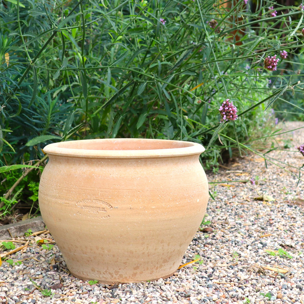 Ein Pfanzentopf, Keramiktopf "Veronica", D=30cm, steht unglasiert und rund auf einem Kiesweg neben hohen Grünpflanzen mit kleinen lila Blüten in einer Gartenanlage.