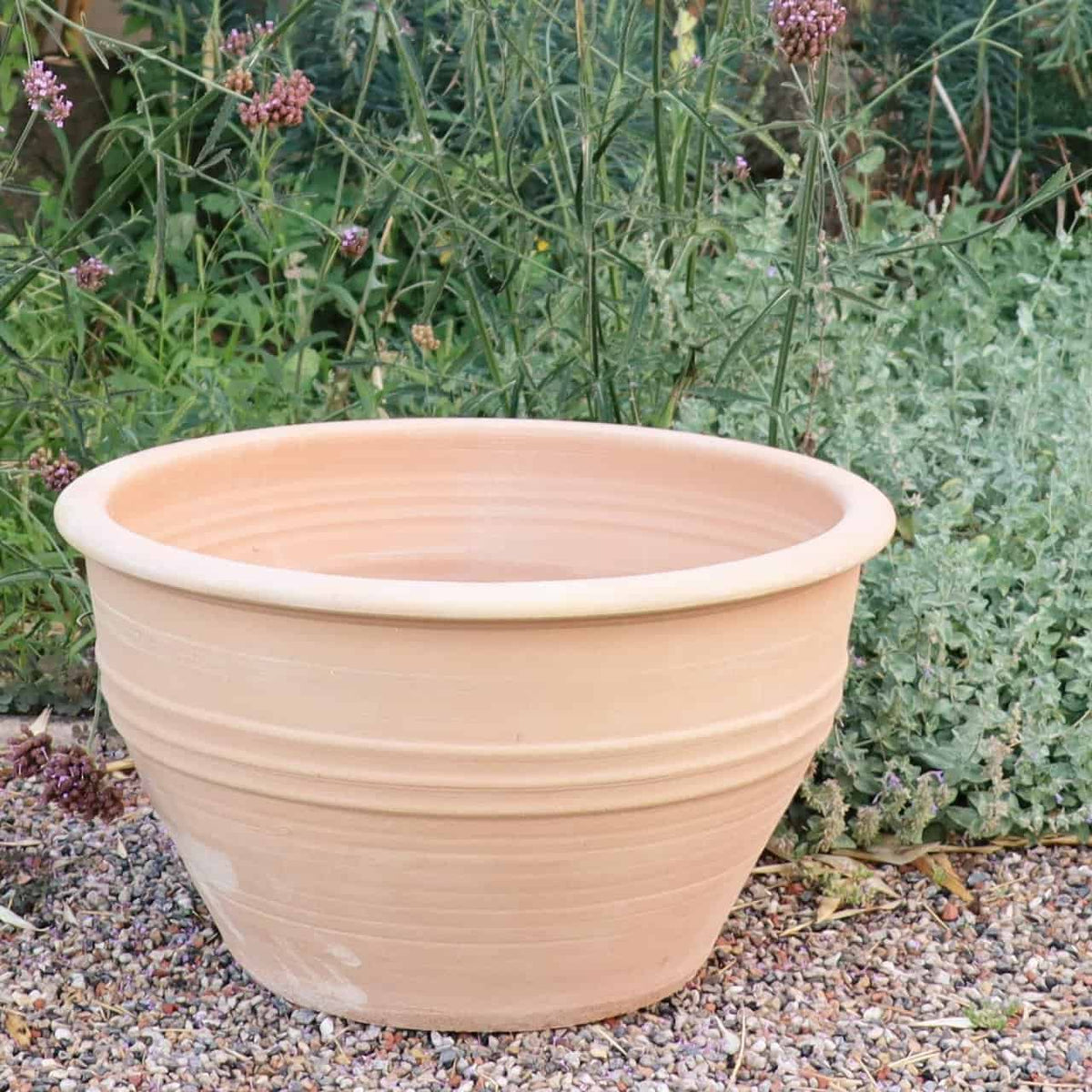 Eine große, leere winterharte Pflanzschale „Veronica“ mit horizontalen Rillen und einer breiten Öffnung von D=55 cm steht auf einer Kiesfläche in einem Garten. Der Hintergrund zeigt dichtes Grün und kleine violette Veronica-Blüten, die die natürliche Außenumgebung ergänzen.