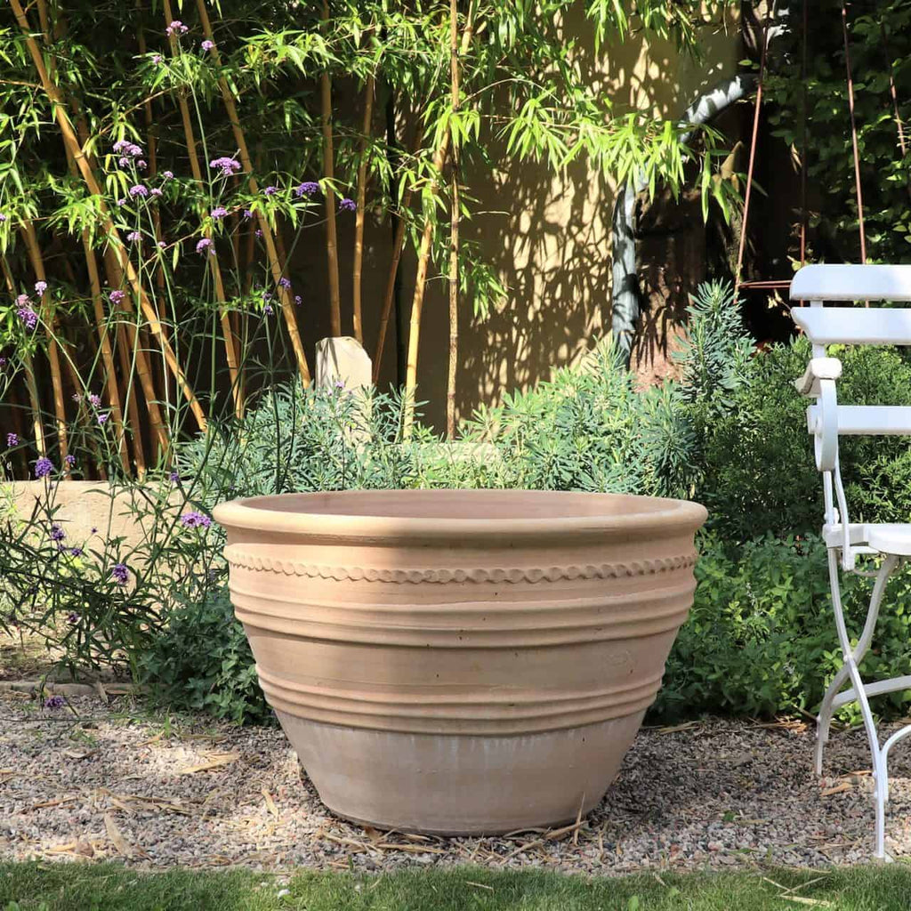 Ein großer frostbeständiger Keramiktopf „Veronica“ aus Terrakotta, D=70 cm, steht auf einem Kiesweg, umgeben von Grünpflanzen und hohem Bambus. Rechts neben dem Pflanzgefäß steht ein weißer Gartenstuhl aus Metall. Die Sonne wirft Schatten und sorgt für eine ruhige Gartenatmosphäre.