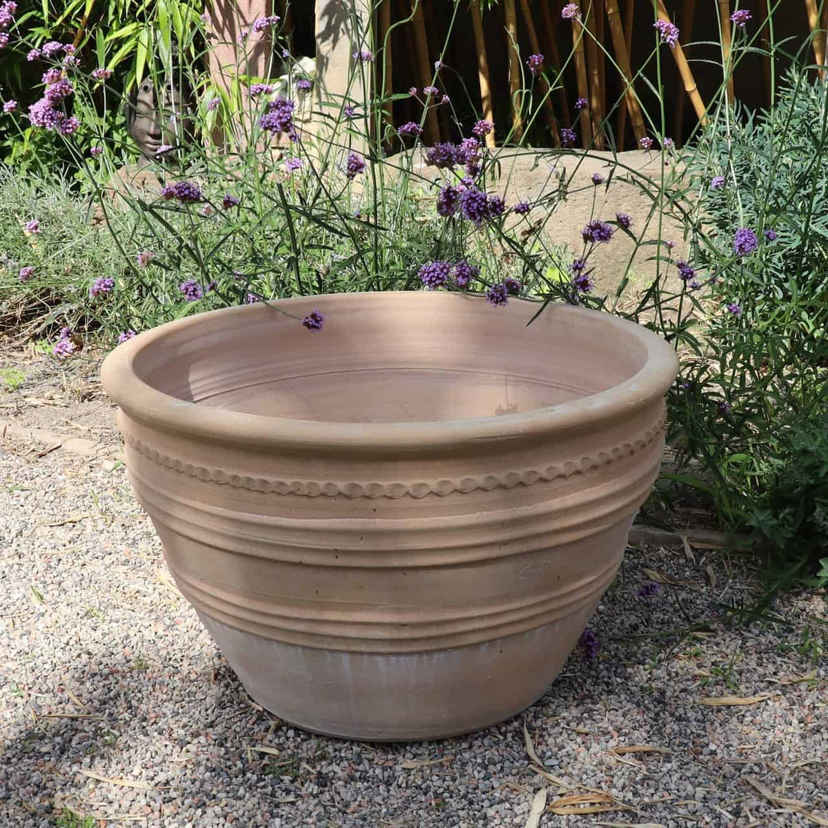 In einem Garten steht ein großer, leerer frostbeständiger Keramiktopf „Veronica“ (D=70cm) aus Terrakotta auf einer Kiesfläche. Um den Topf herum stehen Zweige blühender Pflanzen mit kleinen, violetten Blüten. Sonnenlicht wirft sanfte Schatten und hebt die Textur und Verzierungen dieses schönen Keramiktopfs hervor.