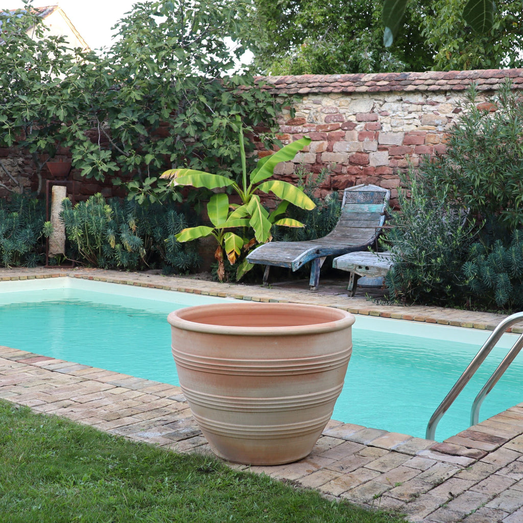 Ein Pool im Hinterhof mit hellblauem Wasser ist von Ziegelsteinen und Grün umgeben, darunter ein Bananenbaum und verschiedene Büsche. Im Vordergrund steht ein großer leerer frostbeständiger Keramiktopf „Veronica“ D=75 cm aus Terrakotta, und in der Nähe der Steinmauer im Hintergrund stehen zwei Holzliegestühle.