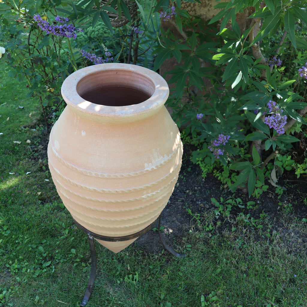 Ein großer, hellbrauner Keramiktopf mit dem Namen „Große liegende Spitzamphore ‚Vitex‘“ mit geriffelten Details steht auf einem schwarzen Metallständer in einem Garten. Der 1,1 Meter hohe Topf ist leer und von üppigem grünem Laub und blühendem Vitex umgeben. Die Szene ist friedlich und gepflegt, mit Gras und kleinen Pflanzen, die rundherum wachsen.