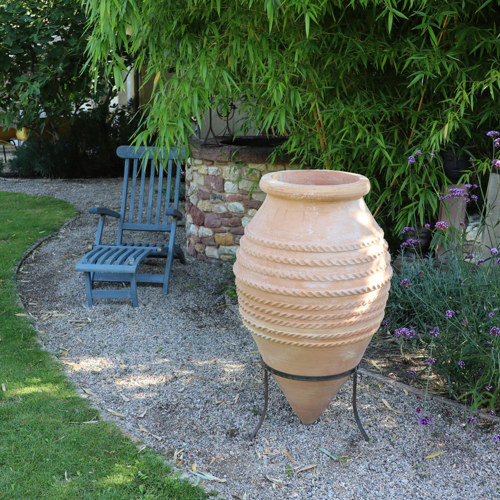Eine große, strukturierte Terrakottavase, die große liegende Spitzamphore „Vitex“ mit einer Höhe von 1,1 Metern, steht auf einem Metallständer inmitten eines Kieswegs in einem Garten. Im Hintergrund steht ein blauer Holzstuhl neben einer Steinmauer, und üppiges Grün, darunter Vitex und blühende Pflanzen, umgibt die Szene.