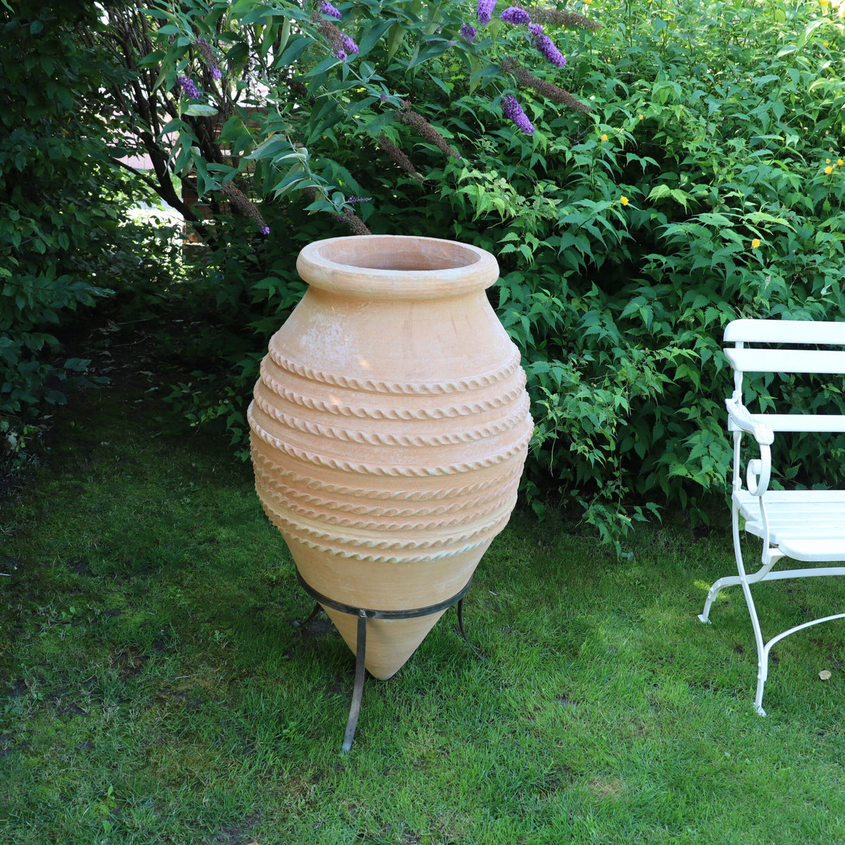 Ein großer, runder Terrakottatopf mit Spiralverzierungen, bekannt als „große liegende Spitzamphore ‚Vitex‘“, steht auf einem Metallständer in einem üppig grünen Garten neben einem weißen Metallstuhl. Im Hintergrund sind dichtes Laub und blühende violette Vitex-Blüten zu sehen.