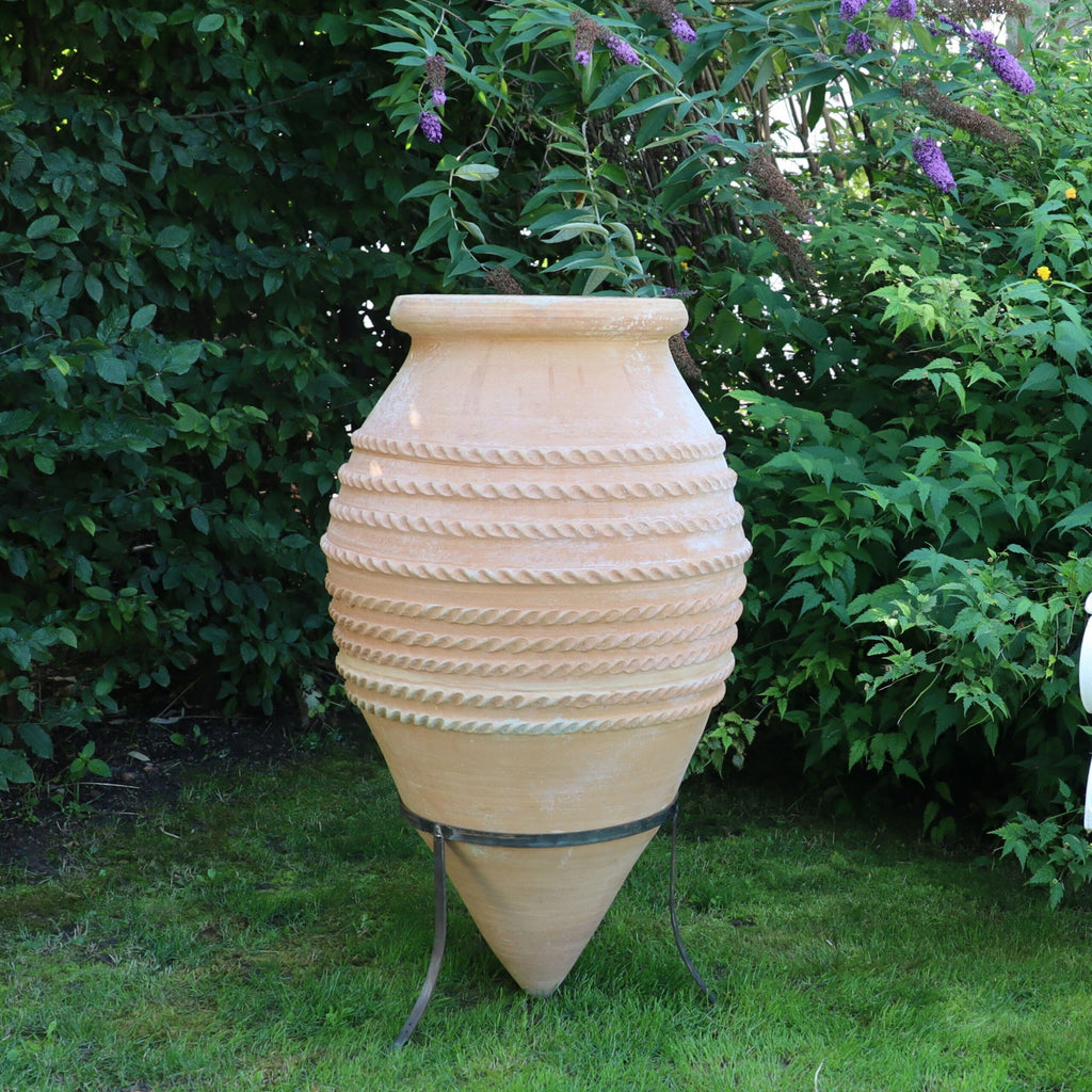 Eine hohe, beige liegende Spitzamphore „Vitex“ aus Keramik steht anmutig auf einem Metallständer in einem Garten, wird teilweise von überhängenden Vitex-Pflanzen beschattet und ist von üppigem Grün umgeben, was eine heitere und natürliche Umgebung schafft.
