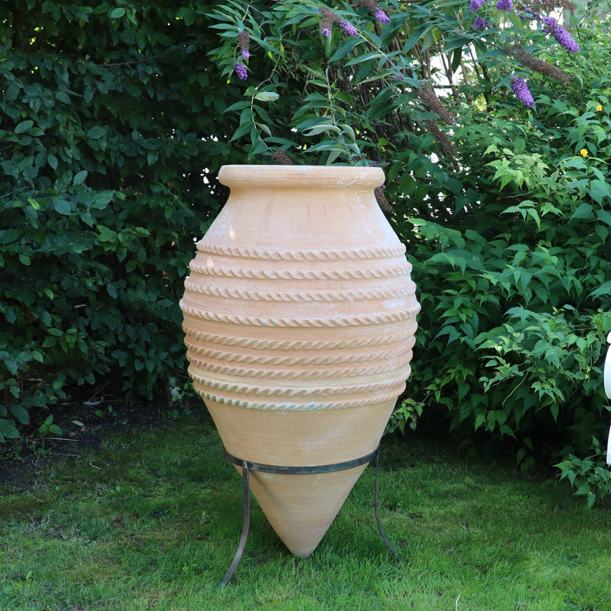 Eine hohe, beige liegende Spitzamphore „Vitex“ aus Keramik steht anmutig auf einem Metallständer in einem Garten, wird teilweise von überhängenden Vitex-Pflanzen beschattet und ist von üppigem Grün umgeben, was eine heitere und natürliche Umgebung schafft.