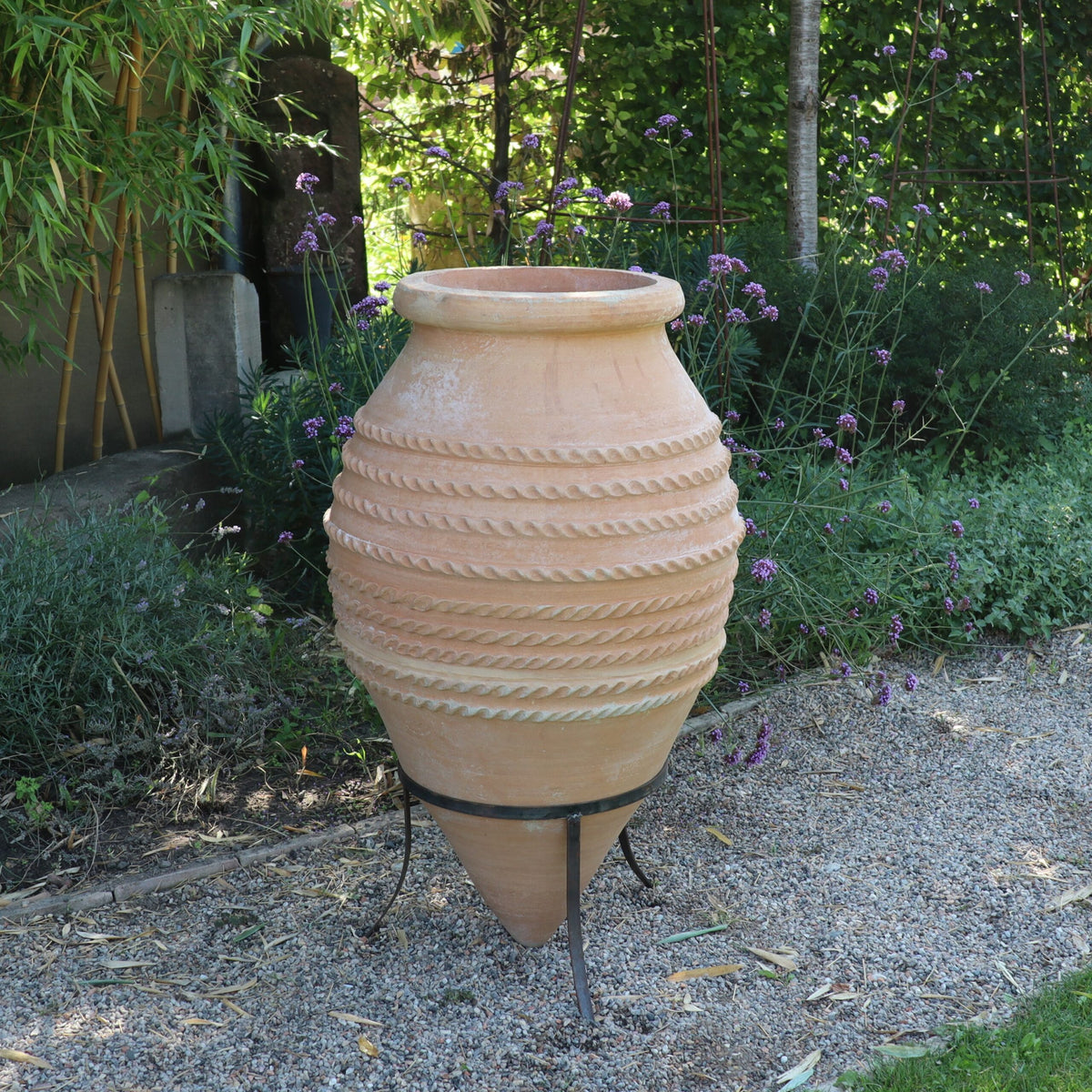 Eine große liegende Amphore „Vitex“ aus Steingut mit strukturiertem Spiralmuster steht auf einem Metallständer in einem Garten. Die Umgebung umfasst grünes Laub, violette Vitex-Blüten und einen Kiesweg.