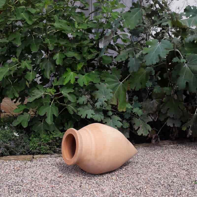 Eine große, hellbraune liegende Spitzamphore „Vitex 2“, H=60cm, liegt seitlich auf einer Kiesfläche vor üppigem grünem Blattwerk. Der konische Topf mit weit geöffneter Mündung wird durch die umgebenden Vitex 2 Blätter perfekt ergänzt.