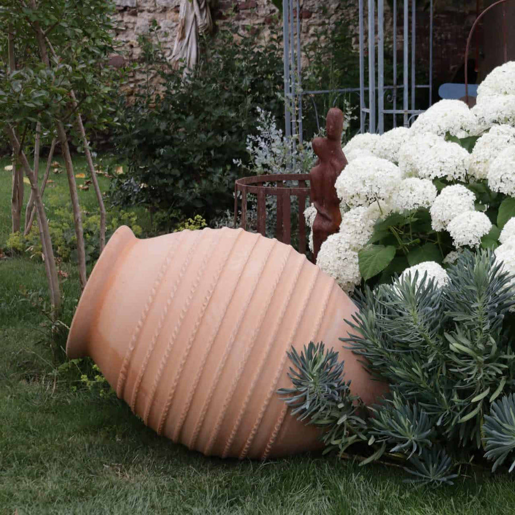 Eine große Terrakotta-Amphore mit dem Namen „Große liegende Amphore ‚Vitex‘“ und einer Höhe von 1,1 Metern liegt auf der Seite in einem üppigen Garten und ähnelt einem antiken Artefakt. Weiße Hortensien blühen dicht um den Topf herum, neben grünem Laub und kleinen Sträuchern wie Vitex, die bis zu 1 Meter hoch werden. Der Garten umfasst verschiedene Pflanzen und Strukturen, die eine heitere und natürliche Atmosphäre schaffen.