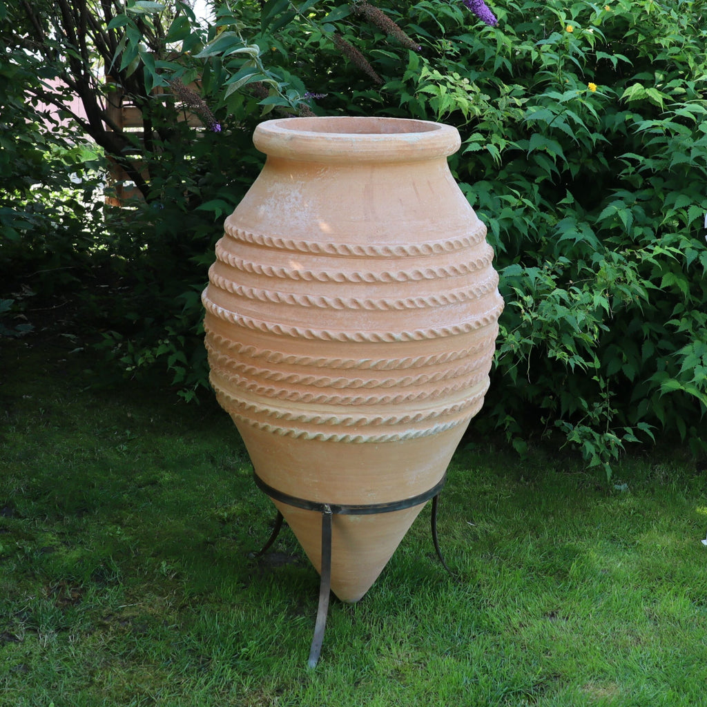 Eine große, beige Keramik-Spitzamphore aus „Vitex“ mit seilartigen Rillen um den Körper steht auf einem Metallständer. Der Topf ist an der Basis konisch und steht auf einem Stück grünem Gras, umgeben von dichtem, belaubtem Gebüsch.