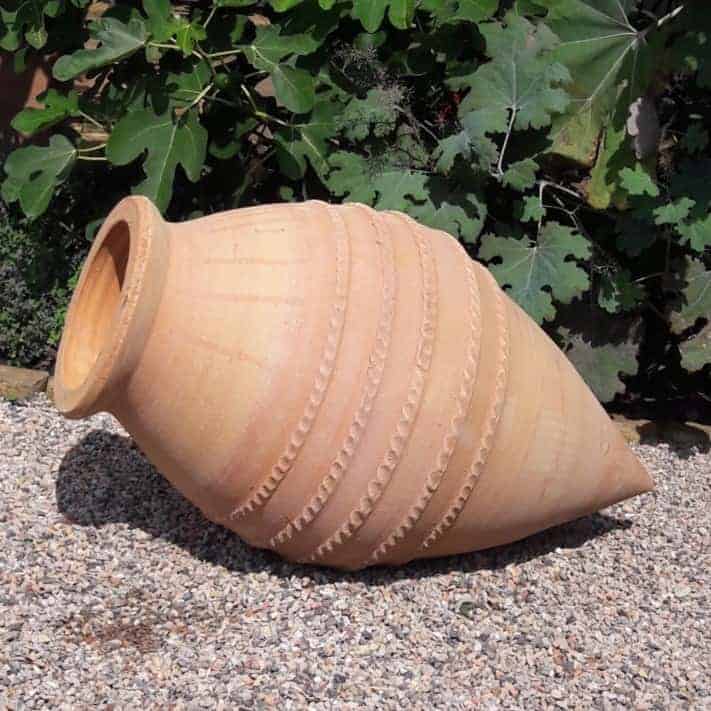 Die handgefertigte liegende Amphore „Vitex“ mit horizontalen Rillen liegt seitlich auf einer Kiesfläche. Der Terrakottatopf hat einen schmalen Boden, der sich zur Mündung hin leicht verbreitert. Im Hintergrund sind grüne Blattpflanzen, darunter Vitex, zu sehen. H=70cm.