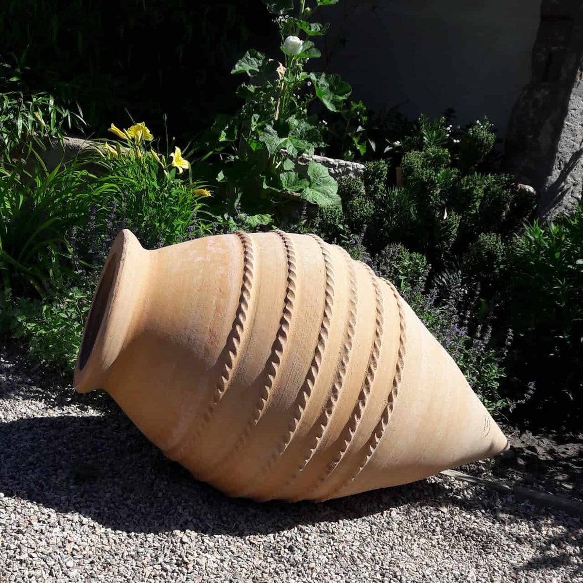 Eine große winterharte liegende Amphore „Vitex“ H=80cm, mit horizontalen seilartigen Mustern und verwitterter Terrakotta-Oberfläche, liegt auf der Seite auf einem Kiesweg. Sie ist von üppigem Grün und verschiedenen blühenden Pflanzen umgeben, wodurch ihr rustikales Aussehen einen Kontrast zur lebendigen Gartenumgebung bildet.