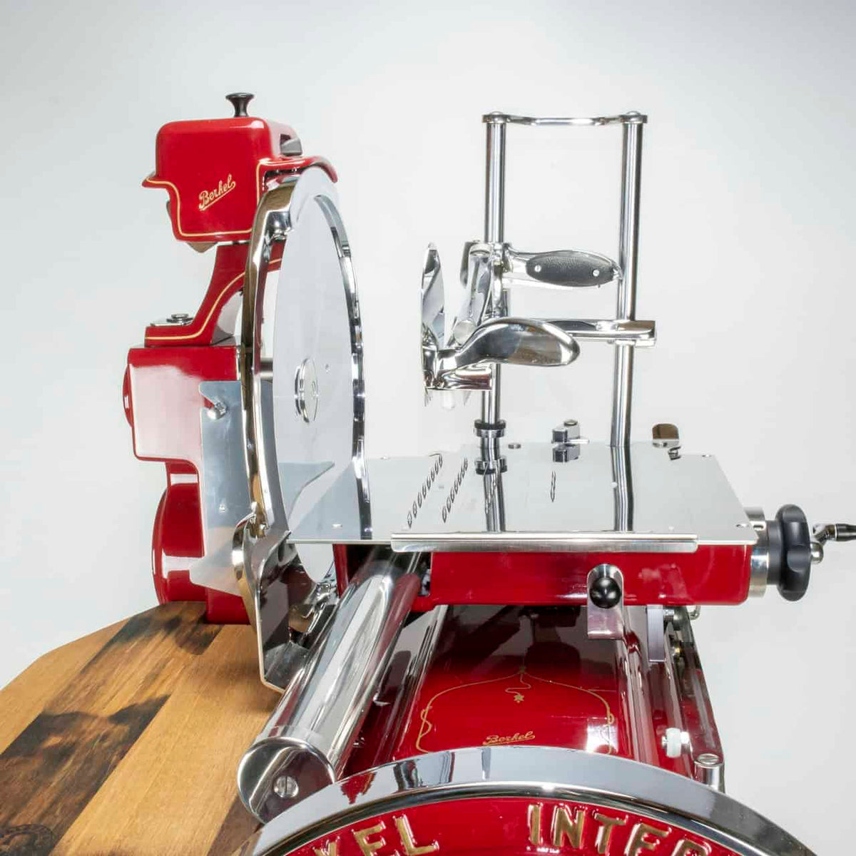 Eine elegante, rote Aufschnittmaschine im Vintage-Stil, die Berkel Volano B114 Rot manuelle Aufschnittmaschine mit passgenauen Einlegebrett, mit Chromakzenten und einem runden Messer steht auf einer Holzoberfläche. Diese Maschine verfügt über einen Griff für die manuelle Bedienung und ein Einlegebrett zum Halten des Fleisches. Der schlichte weiße Hintergrund unterstreicht das Design der Maschine.