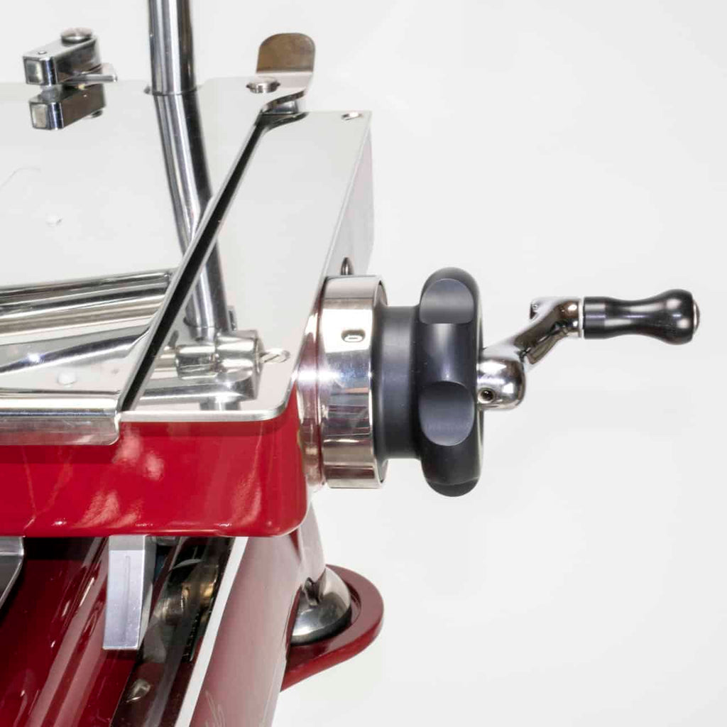 Nahaufnahme einer manuellen Aufschnittmaschine Berkel Volano B114 Rot mit passgenauem Einlegebrettgriff. Der Griff aus schwarzem Kunststoff und Metallkomponenten ist an einer roten Basis befestigt. Die scharfe Klinge und die glänzenden Metalloberflächen der Maschine sind vor einem schlichten weißen Hintergrund sichtbar.