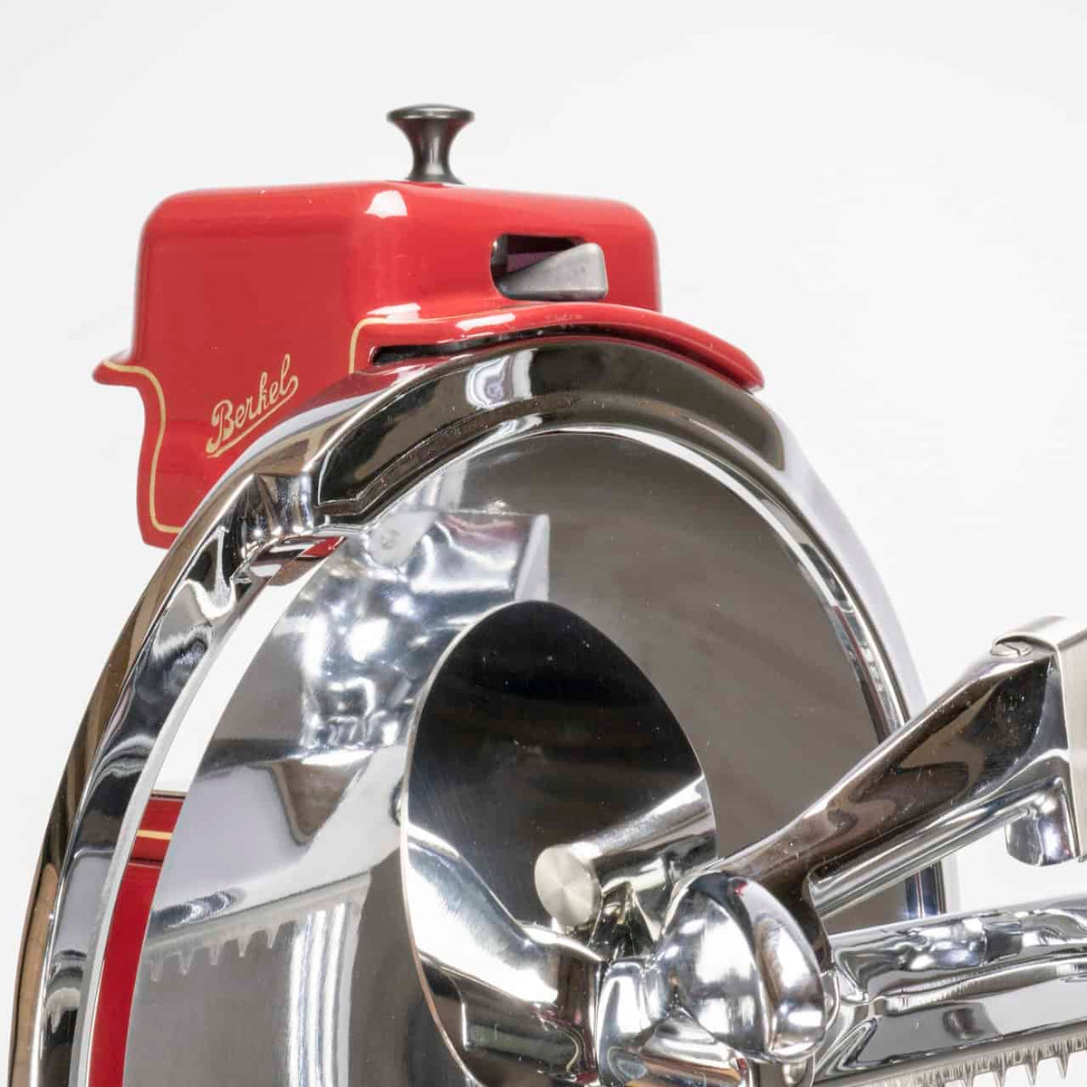 Eine Nahaufnahme der glänzenden, verchromten manuellen Aufschnittmaschine Berkel Volano B114 Rot mit passgenauem Einlegebrett zeigt ihr Vintage-Fleischschneidemaschinen-Design mit einer roten Oberseite mit der Aufschrift „Berkel Volano B114“. Das Bild hebt die Klinge und den Griff der Schneidemaschine hervor und reflektiert das Licht von der Metalloberfläche. Der schlichte weiße Hintergrund betont die präzisen Designdetails dieser manuellen Fleischschneidemaschine.