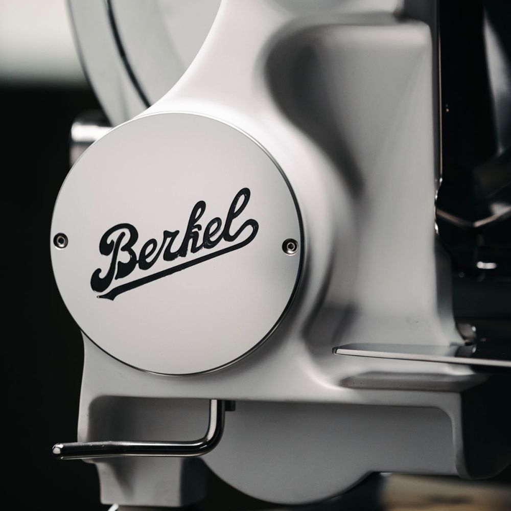 Nahaufnahme einer weißen Berkel Volano B114X - Exklusive Schwungradmaschine mit Standfuß und Fassholzbrett, auf der das kursive "Berkel"-Logo auf einer runden Metallplatte zu sehen ist.