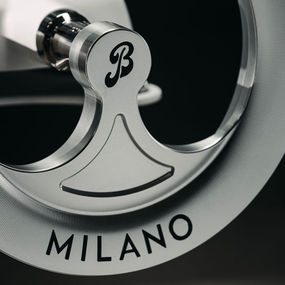 Nahaufnahme des Metallrads der Berkel Volano B114X - Exklusive Schwungradmaschine mit Standfuß und Fassholzbrett. Das kursive "B" in der Mitte und der eingravierte Schriftzug "MILANO" am äußeren Rand unterstreichen das schlanke, präzise Design.