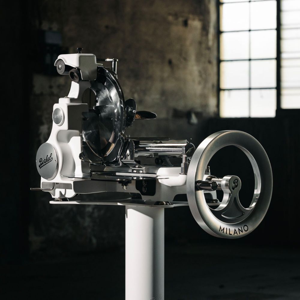 A Berkel Volano B114X - Exklusive Schwungradmaschine mit Standfuß und Fassholzbrett steht in einem schwach beleuchteten industriellen Raum mit großen Fenstern im Hintergrund.