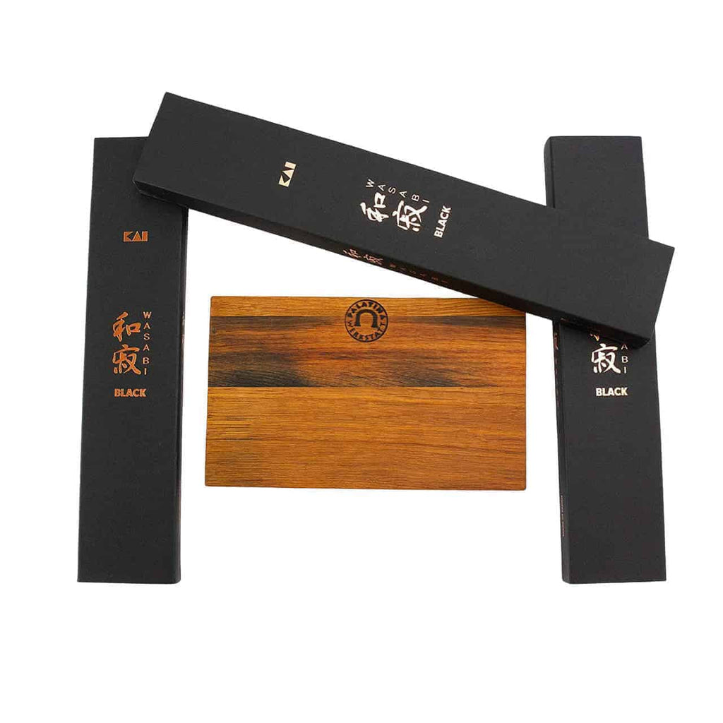 Ein Schneidebrett aus Fassholz wird neben drei schwarzen rechteckigen Schachteln mit goldenem und weißem Text ausgestellt. In der Mitte des Schneidebretts befindet sich ein Markenlogo. Auf den Schachteln steht „Kai Wasabi Black“, was auf die Marke und insbesondere auf die im Kai Wasabi Black Kochmesser-Set enthaltenen Produkte hinweist: ein 20 cm langes Kochmesser, ein 15 cm langes Allzweckmesser und ein 10 cm langes Officemesser.