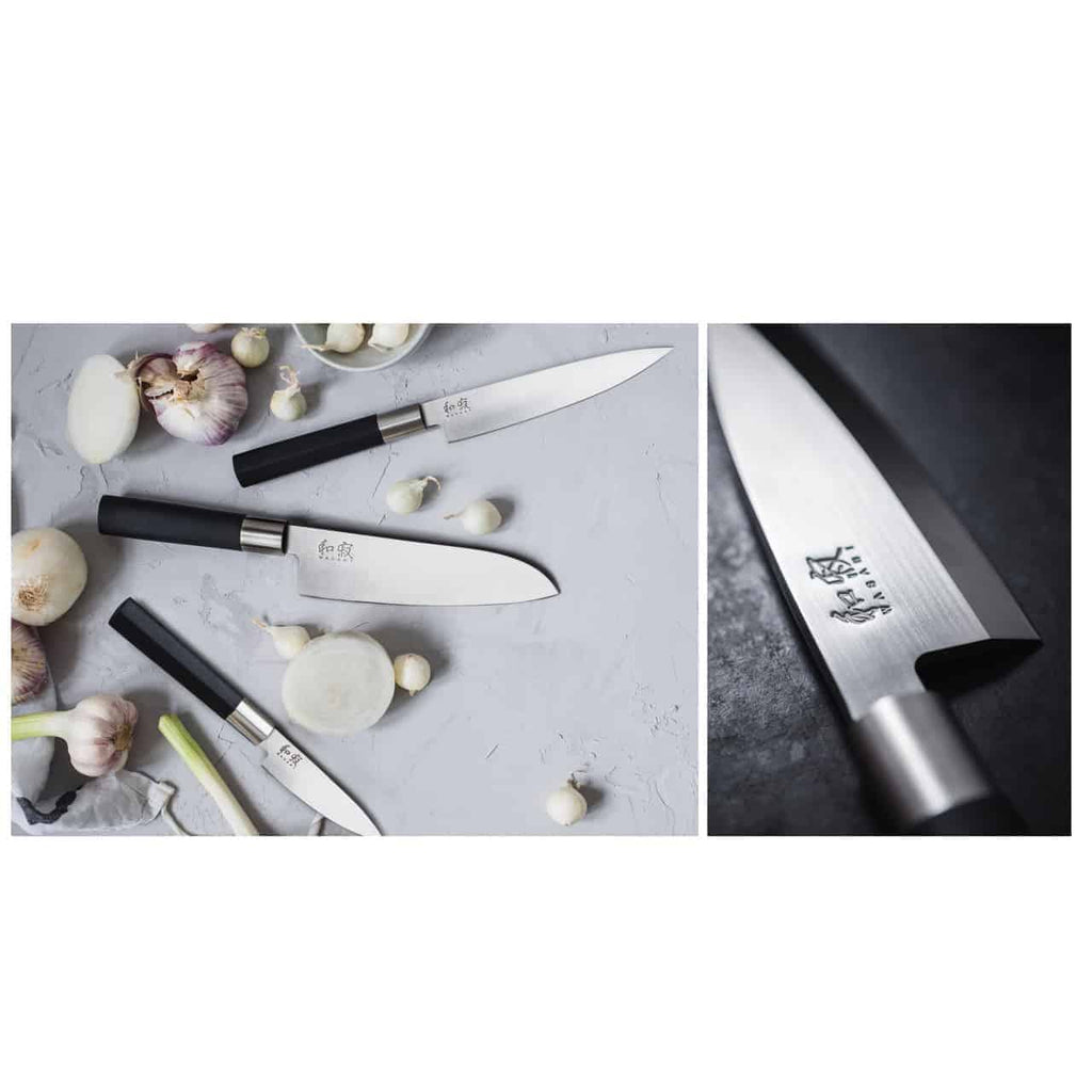 Das Kai Wasabi Black Messerset umfasst ein 20 cm langes Kochmesser, ein 15 cm langes Allzweckmesser und ein 10 cm langes Schälmesser, alle mit schwarzen Griffen und japanischen Inschriften auf den Klingen. Die Messer werden auf einer grauen Oberfläche mit Knoblauchknollen und -zehen präsentiert. Das Bild ist geteilt; die rechte Seite zeigt eine Nahaufnahme der Klinge eines Messers vor dem mitgelieferten Schneidebrett aus Fassholz, 25 x 15 cm groß.