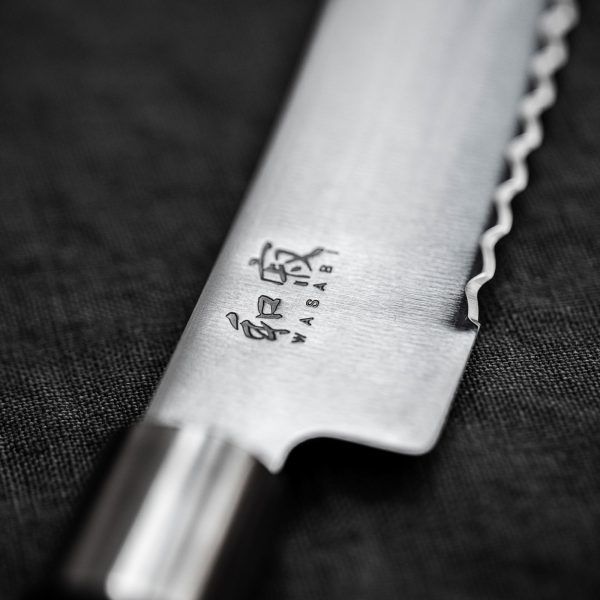 Nahaufnahme eines silbernen Wellenschliffmessers mit japanischen Schriftzeichen und dem Wort „WASABI“, das in die Klinge eingraviert ist. Das Messer, Teil des KAI Weihnachtsangebots 4-teiliges Geschenkset – Wasabi Black – limitiertes 1.000 Stk. Set, ruht auf einer dunkel gefärbten Stoffoberfläche. Die Klinge zeigt feine Handwerkskunst mit einer gewellten, gezackten Kante, perfekt für jede kulinarische Aufgabe.