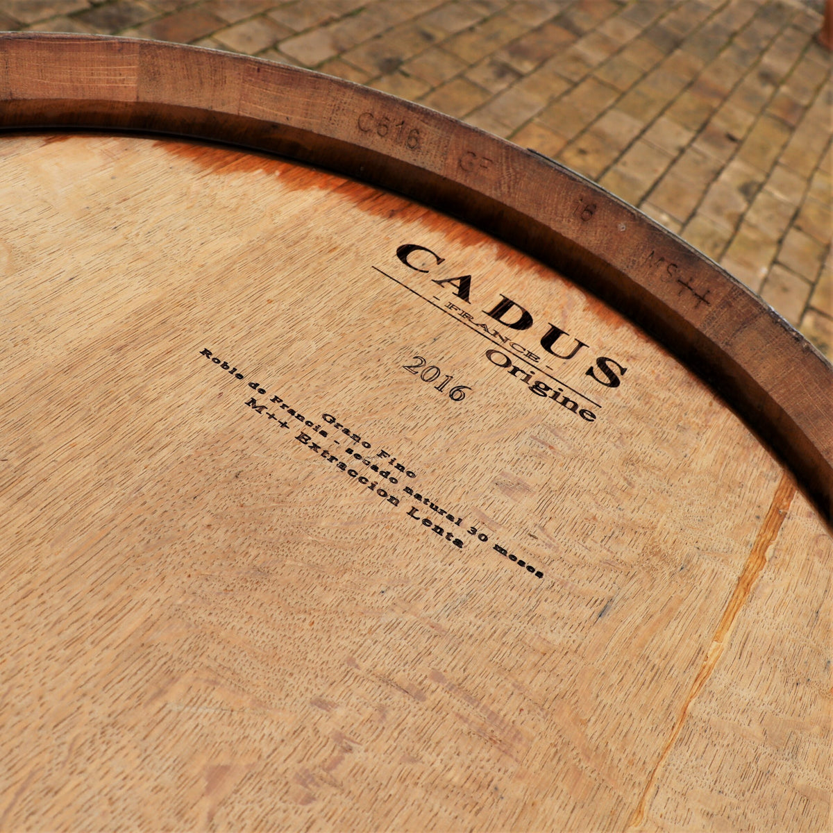 Eine Nahaufnahme eines originalen 300-Liter-Weinfasses, Barriquefasses, Regenfasses aus Eiche, gebraucht mit der Aufschrift „Cadus Origine 2016“ und einem zusätzlichen Text auf Französisch, das im Freien auf einem Kopfsteinpflaster steht. Das Fass hat eine reiche, natürliche Holzstruktur und ein Eisenband um den oberen Rand.