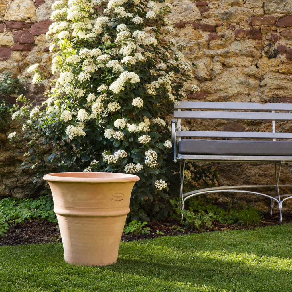Das winterharte Terracotta-Pflanzgefäß "Yucca", H=35 cm, steht auf einer grünen Wiese neben weiß blühenden Sträuchern und einer Metallbank mit grauen Kissen, vor einer rustikalen Steinmauer.