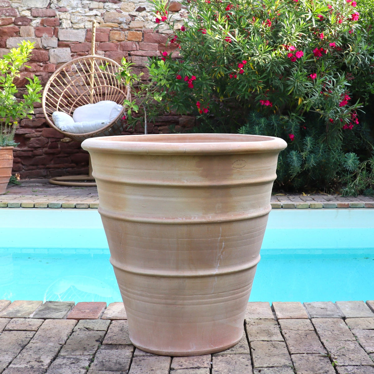 Der winterfeste Terracotta Blumenkübel groß "Yucca", H=60 cm, steht auf Steinplatten am Pool, umgeben von blühenden Büschen, einer Yucca-Pflanze und einem hängenden Korbstuhl im Hintergrund.