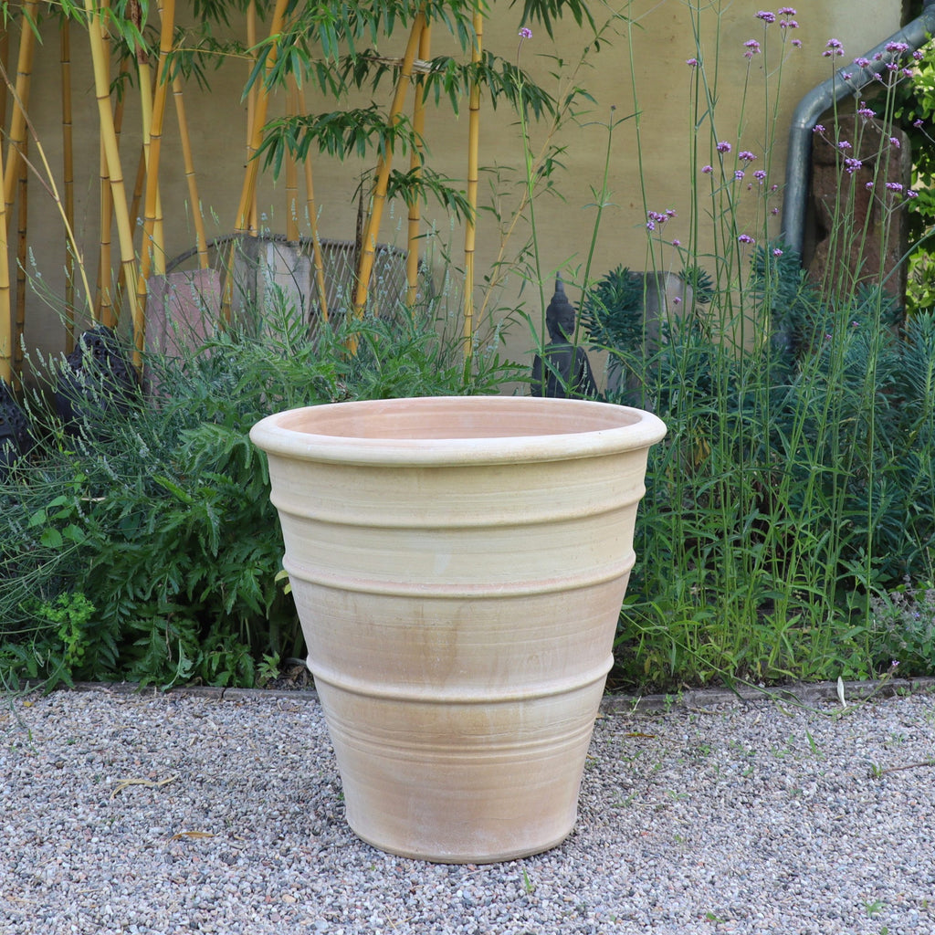 Der winterfeste, terrakottafarbene Blumenkübel groß "Yucca", H=60 cm, steht leer auf Kies zwischen hohen Grünpflanzen, einer markanten Yucca und Bambus vor einer beigen Wand.