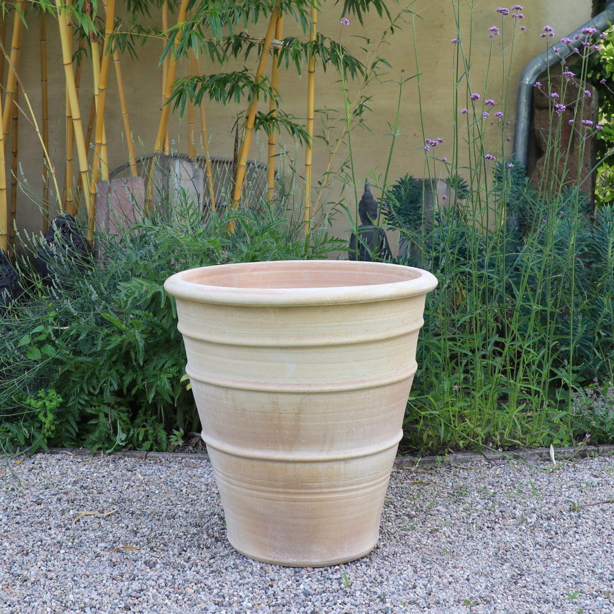 Der winterfeste, terrakottafarbene Blumenkübel groß "Yucca", H=60 cm, steht leer auf Kies zwischen hohen Grünpflanzen, einer markanten Yucca und Bambus vor einer beigen Wand.