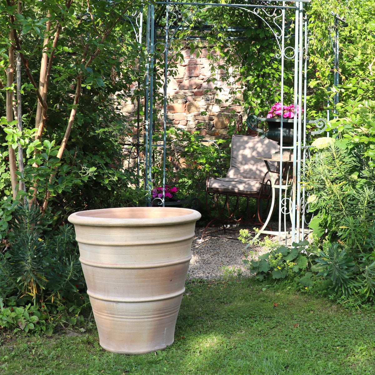 Der winterfeste Terrakotta Blumenkübel groß "Yucca", H=60 cm, steht auf dem Rasen eines üppigen Gartens nahe einem schmiedeeisernen Pavillon mit Stuhl, umgeben von Grün und Blüten - perfekt für Ihre Yucca.