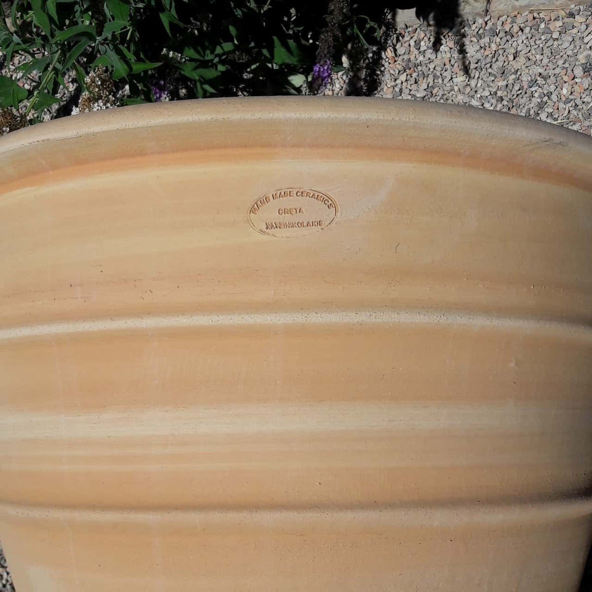 Eine Nahaufnahme eines großen Blumenkübels aus Terrakotta hoch „Yucca“, H=80 cm. Der Topf weist dezente horizontale Streifen und einen ovalen Stempel mit der Aufschrift „Poterie Naas Ceramics, Grez, Ramondorage“ auf. Der umgebende Hintergrund besteht aus grünem Laub und einer Kiesfläche, in deren Nähe teilweise eine Yucca-Pflanze zu sehen ist.