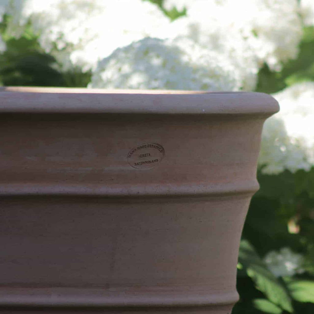 Eine Nahaufnahme eines großen Blumenkübels aus Terrakotta hoch „Yucca“, H=80 cm, vor einem Hintergrund aus weißen Hortensienblüten. Der Topf hat ein geprägtes Etikett, aber der Text ist nicht klar lesbar. Die leuchtend weißen Blüten und grünen Blätter sind im Hintergrund sanft verschwommen und heben die Details des Topfes hervor.