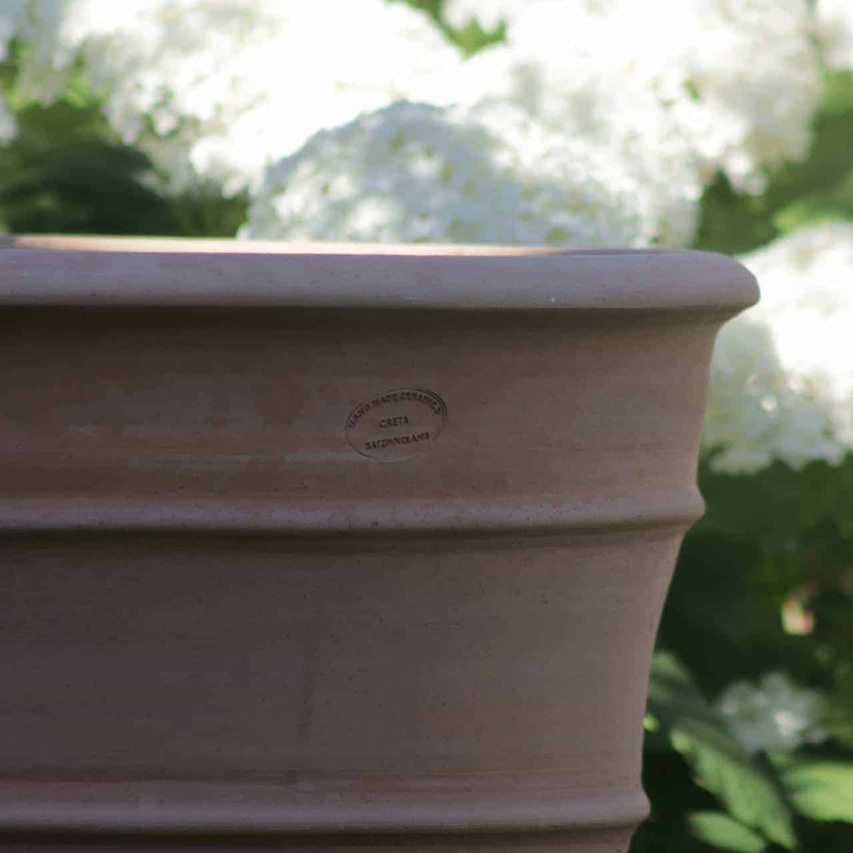 Eine Nahaufnahme eines großen Blumenkübels aus Terrakotta hoch „Yucca“, H=80 cm, vor einem Hintergrund aus weißen Hortensienblüten. Der Topf hat ein geprägtes Etikett, aber der Text ist nicht klar lesbar. Die leuchtend weißen Blüten und grünen Blätter sind im Hintergrund sanft verschwommen und heben die Details des Topfes hervor.