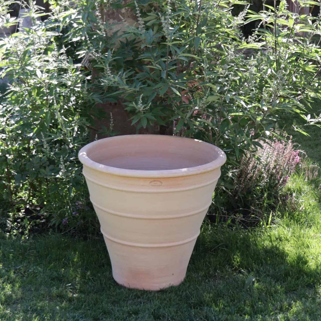 Ein großes, leeres Terrakotta-Pflanzgefäß „Yucca“, 70 cm hoch, steht auf einer Rasenfläche vor einem üppigen grünen Busch. Der Busch ist hoch und belaubt und bietet einen natürlichen Hintergrund für den Topf. Sonnenlicht wirft weiche Schatten und hebt die Texturen der Terrakotta und des Laubes hervor.