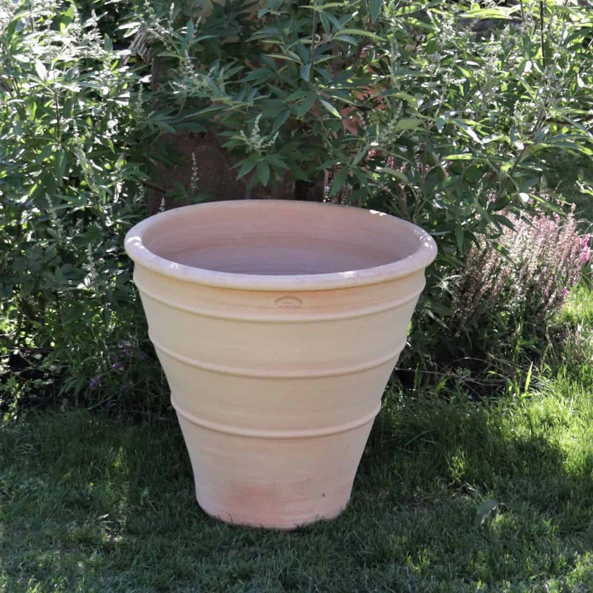 Ein großes beiges Terrakotta-Pflanzgefäß „Yucca“ mit gestuftem Design steht majestätisch auf einem Grasfleck im Garten, umgeben von üppigen grünen Pflanzen und Blättern, und trägt zur ruhigen Landschaft bei.