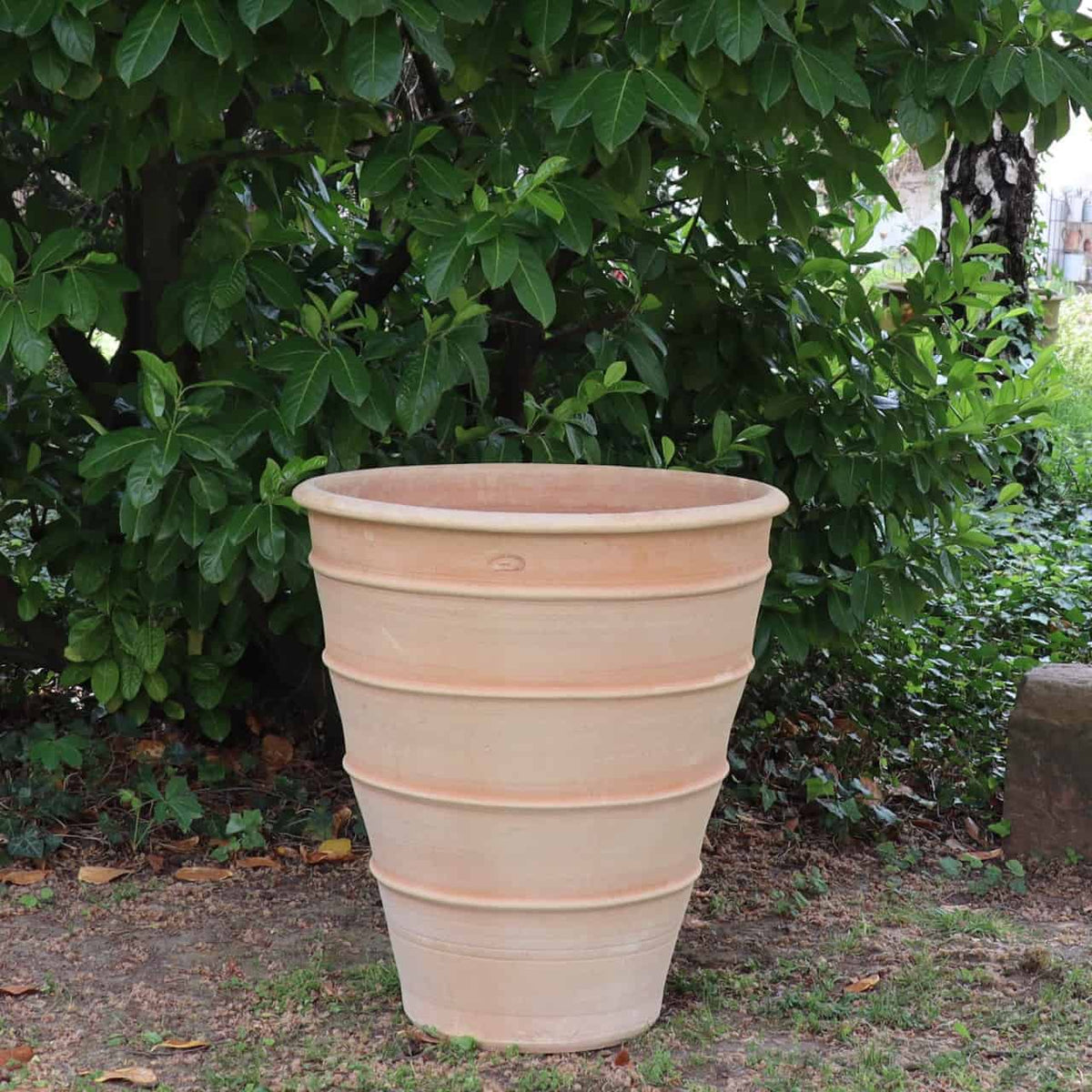 Ein Terrakotta-Pflanzgefäß „Yucca“ mit einer Höhe von 70 cm und einem geriffelten, gestuften Design steht auf dem Boden vor dichtem grünem Laub. Dieser große, leere Topf steht perfekt im Freien in der Natur und ist somit ein idealer Ort für eine Yucca-Pflanze.