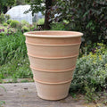 Ein großes, beiges Terrakotta-Pflanzgefäß „Yucca“, 70 cm hoch, mit horizontalen Rillen, schmückt ein Holzdeck in einem üppigen Garten. Eine majestätische Yucca-Pflanze verleiht dem Topf Charakter, während das umgebende Grün und die Bäume im Hintergrund eine ruhige Außenumgebung schaffen.