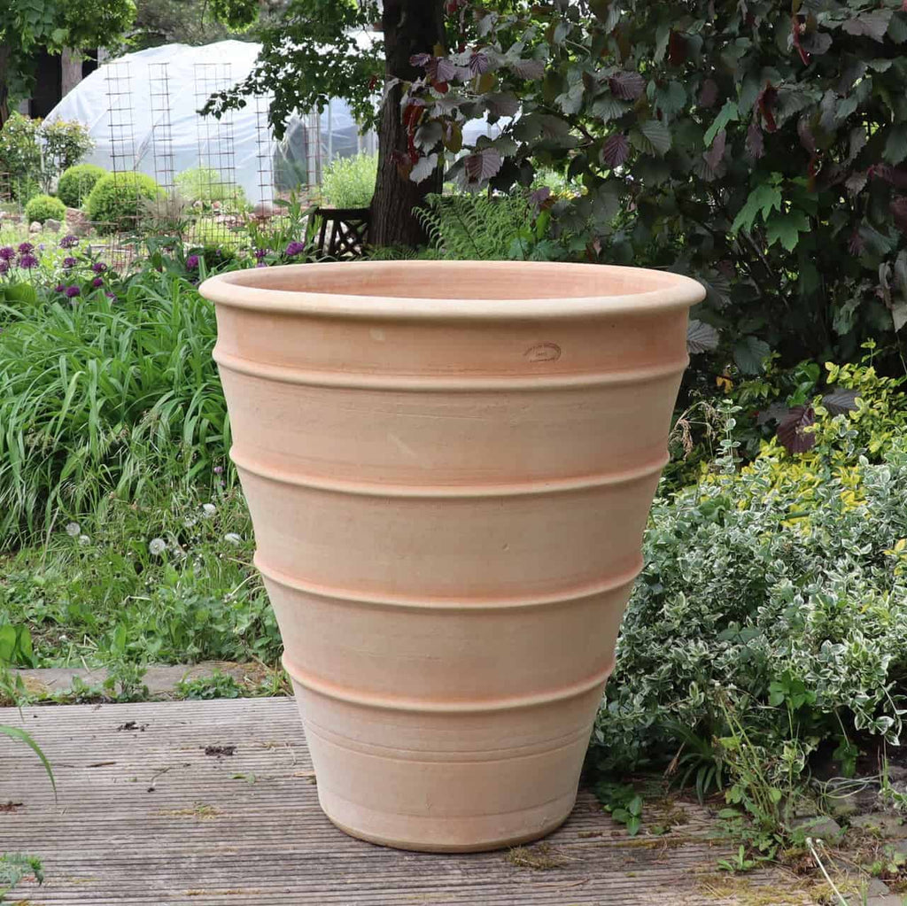 Ein großes, beiges Terrakotta-Pflanzgefäß „Yucca“, 70 cm hoch, mit horizontalen Rillen, schmückt ein Holzdeck in einem üppigen Garten. Eine majestätische Yucca-Pflanze verleiht dem Topf Charakter, während das umgebende Grün und die Bäume im Hintergrund eine ruhige Außenumgebung schaffen.