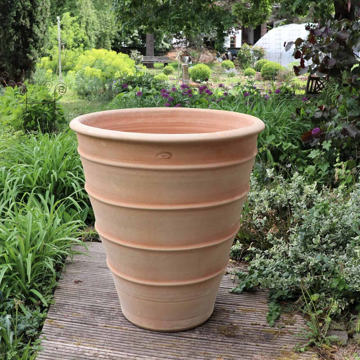 Ein großes, leeres Terrakotta-Pflanzgefäß „Yucca“ mit einer Höhe von 70 cm und geripptem Design steht auf einem Holzweg in einem üppigen Garten. Der Garten, gefüllt mit verschiedenen Grünpflanzen, Blumen und kleinen Sträuchern, schafft eine heitere und lebendige Atmosphäre.