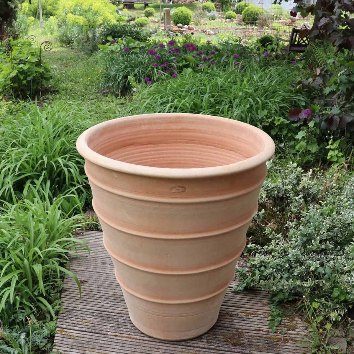 Ein großes, leeres Terrakotta-Pflanzgefäß „Yucca“ mit geripptem Design steht auf einem Holzweg. Es befindet sich in einem üppigen Garten mit verschiedenen Grünpflanzen, darunter einige auffällige Yuccas, Sträucher und violette Blumen im Hintergrund.
