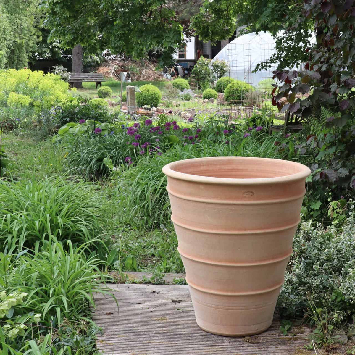 Ein großes, leeres Terrakotta-Pflanzgefäß „Yucca“ mit einer Höhe von 70 cm ziert einen Holzweg, der durch einen üppigen, grünen Garten mit verschiedenen Pflanzen und Büschen führt. Im Hintergrund ist dichtes Laub zu sehen und man sieht ein Haus. Es ist ein heller und sonniger Tag.