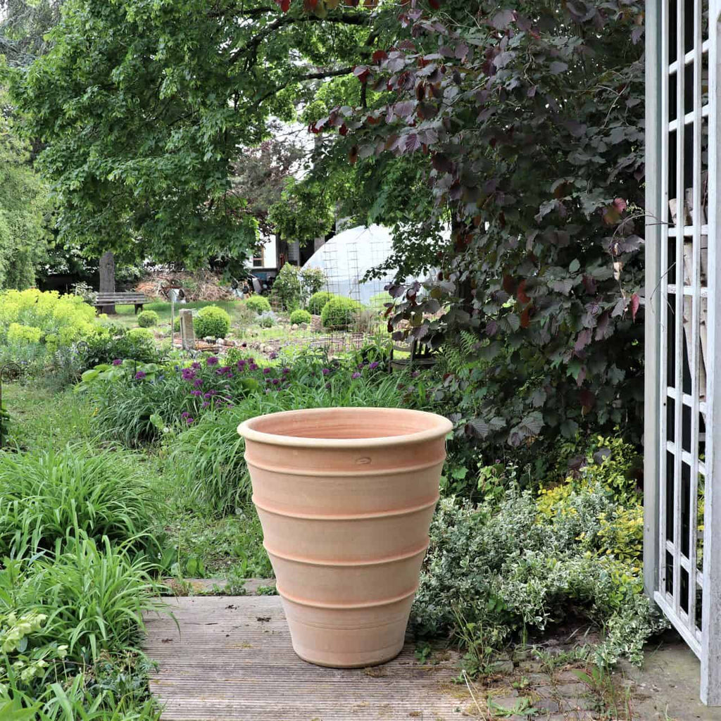 Ein großer Blumenkübel „Yucca“ aus Terrakotta mit einer Höhe von 80 cm ziert ein Holzdeck in einem üppigen Garten. Um den Topf herum stehen verschiedene Grünpflanzen, darunter Sträucher und Bäume. Im Hintergrund sind ein Gewächshaus und weitere Grünpflanzen zu sehen, die für eine friedliche, ländliche Atmosphäre sorgen.