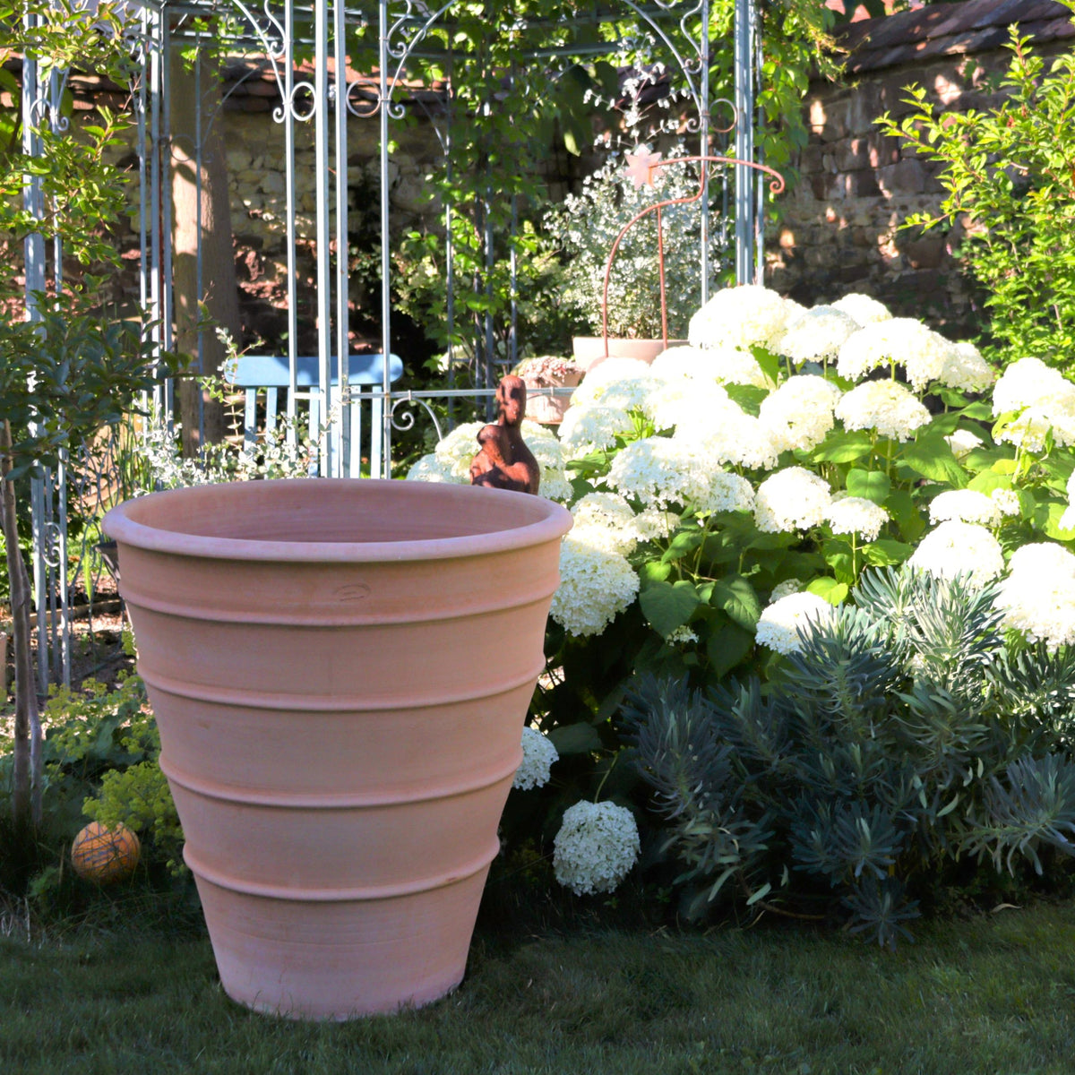 Ein großer Blumenkübel hoch „Yucca“ (H=80 cm) aus Terrakotta steht in einem üppigen Garten mit weißen Hortensien und Grünpflanzen, während ein Metallpavillon und eine kleine Hundeskulptur im gesprenkelten Sonnenlicht hervorstechen.