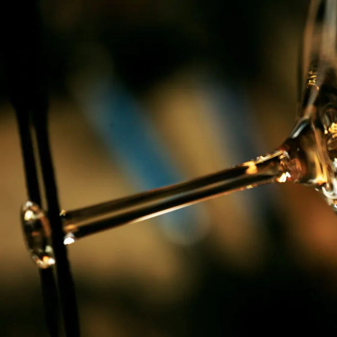 Nahaufnahme des Stiels des Zaltoglas Denk'Art mundgeblasenes Gravitas Omega, möglicherweise von einem Weinglas, vor einem verschwommenen dunklen und goldenen Hintergrund. Das Bild hebt die elegante Kurve und die Reflexion dieses handgefertigten Glases hervor.