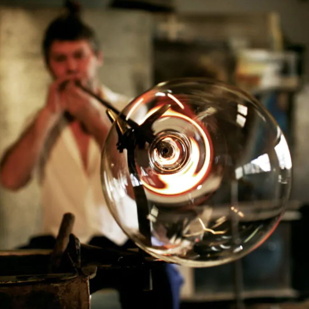 Eine Person bläst in ein Metallrohr, um eine große, klare Zaltoglas Denk'Art mundgeblasenes Gravitas Omega-Blase in einer Glasbläserwerkstatt zu formen. Das sich ausdehnende Glas ist im Fokus, der Handwerker ist im Hintergrund verschwommen.