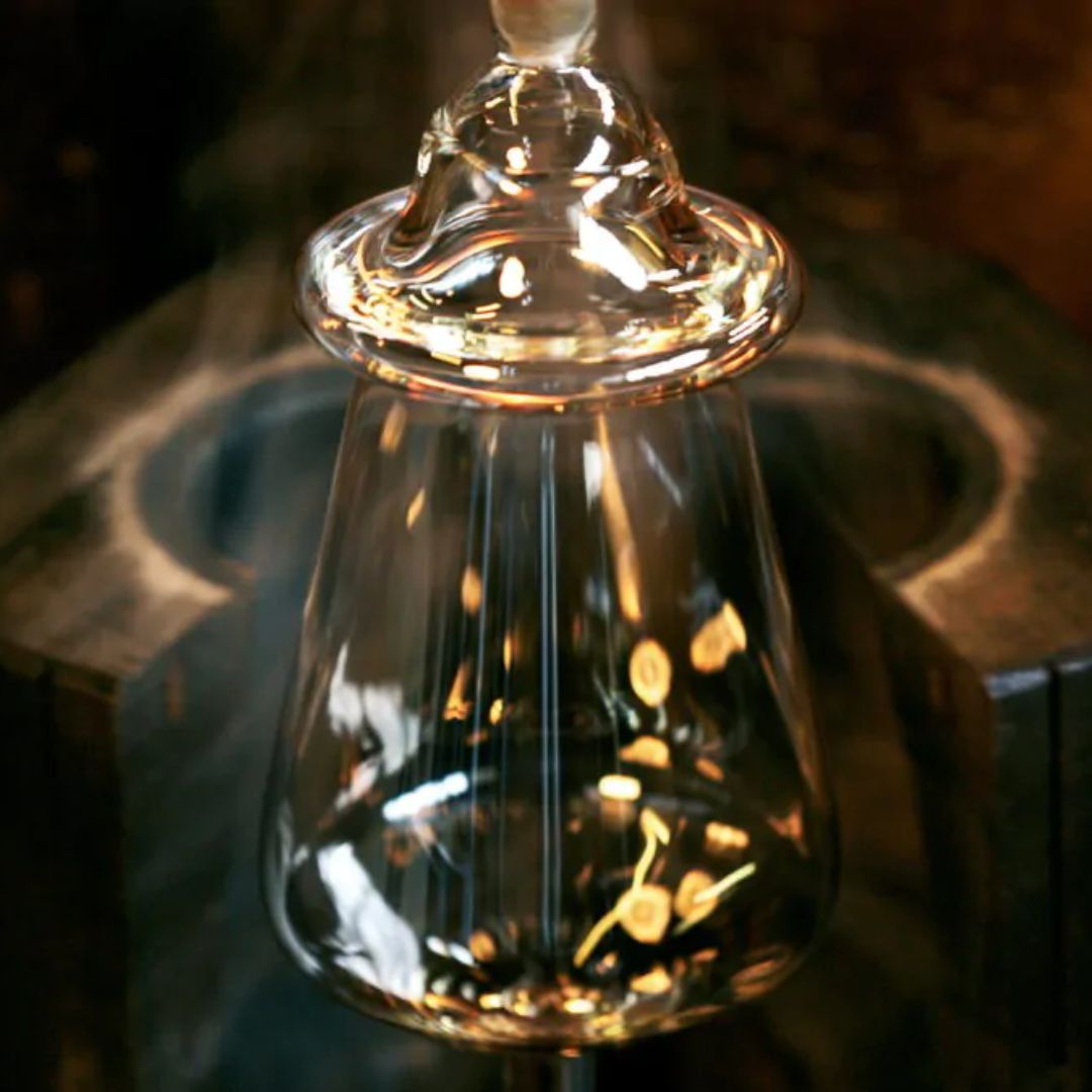 Eine Nahaufnahme des Zaltoglas Denk'Art mundgeblasener Karaffe Nr. 150, der während des Glasblasens geformt wird, wobei sich das Licht auf seiner transparenten Oberfläche vor einem dunklen, unscharfen Hintergrund spiegelt.