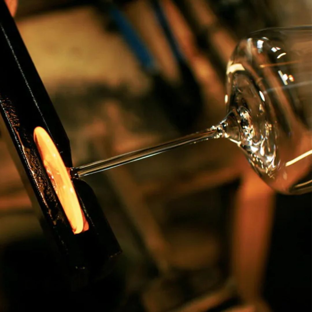 Eine Nahaufnahme zeigt einen leuchtend orangefarbenen Klecks geschmolzenen Glases, der bei der Herstellung des Zaltoglas Denk'Art mundgeblasener Karaffe Nr. 150 mit einer schwarzen Metallzange geformt und befestigt wird.