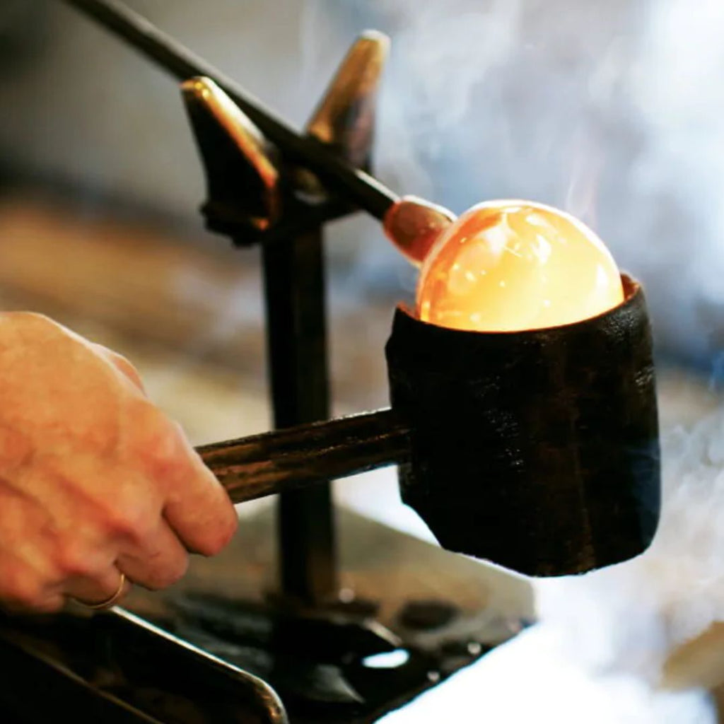Eine Person formt den Zaltoglas Denk'Art mundgeblasener Karaffe Nr. 150 mit einer Metallzange und einer Form, während beim traditionellen Glasblasverfahren Dampf aufsteigt.